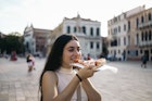 Young woman eating a piece of pizza in a city square. She smiles enjoying her food in Italy

