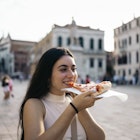 Young woman eating a piece of pizza in a city square. She smiles enjoying her food in Italy

