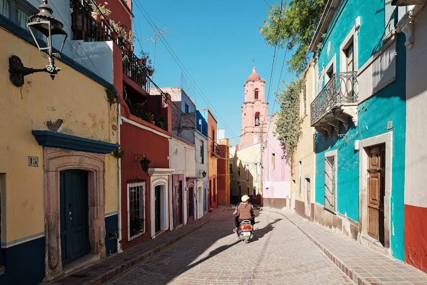 2 people leave on a motorbike on a cobblestone road with colorful homes on either side. The historic Town of Guanajuato is listed as a UNESCO World Heritage Site