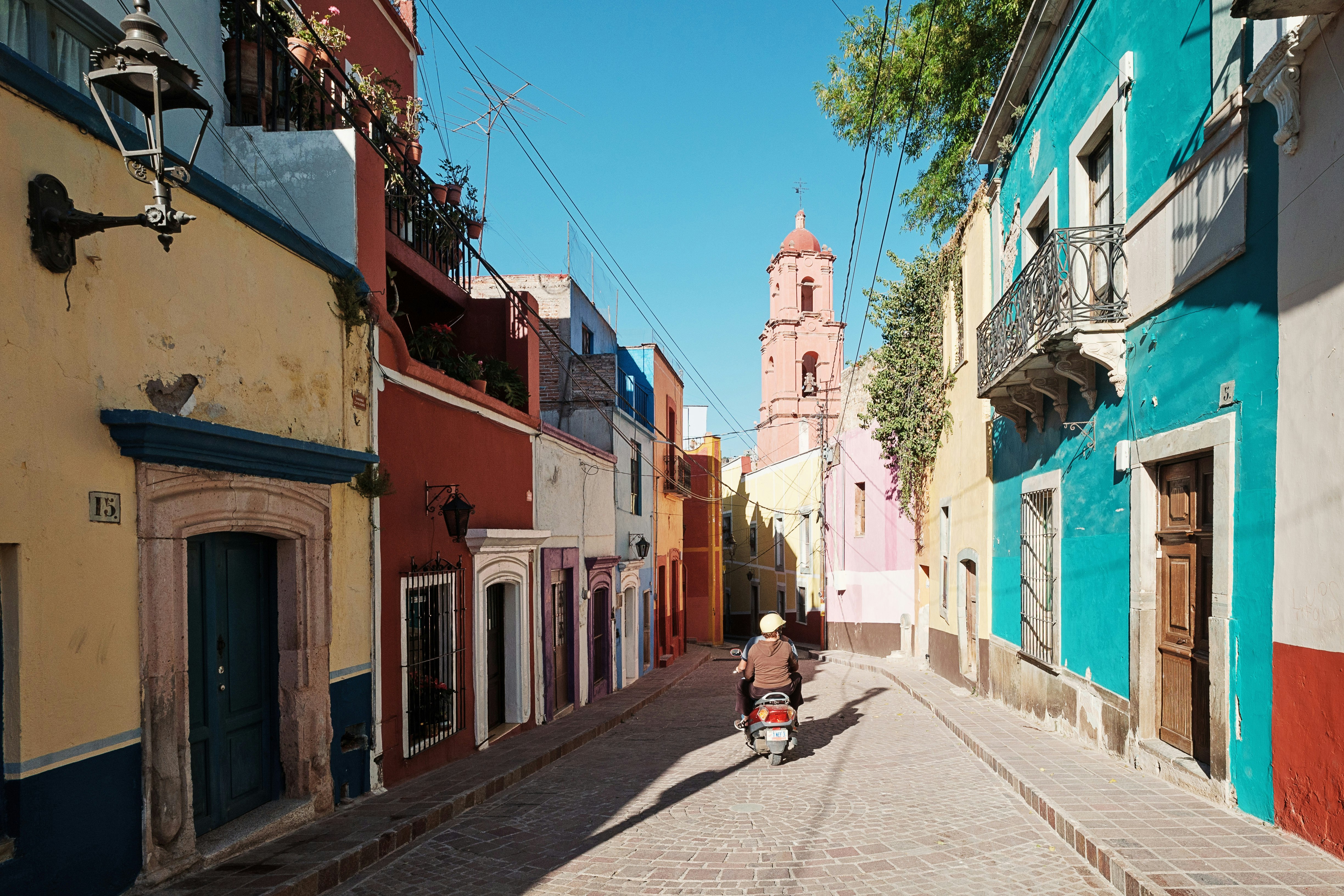 2 people leave on a motorbike on a cobblestone road with colorful homes on either side. The historic Town of Guanajuato is listed as a UNESCO World Heritage Site
