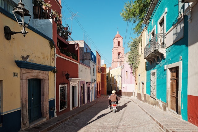 2 people leave on a motorbike on a cobblestone road with colorful homes on either side. The historic Town of Guanajuato is listed as a UNESCO World Heritage Site