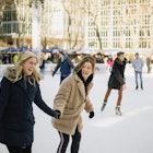 A front-view shot of two mid-adult women ice skating together in an ice rink in New York City, they are wearing warm clothing, holding hands and laughing together.