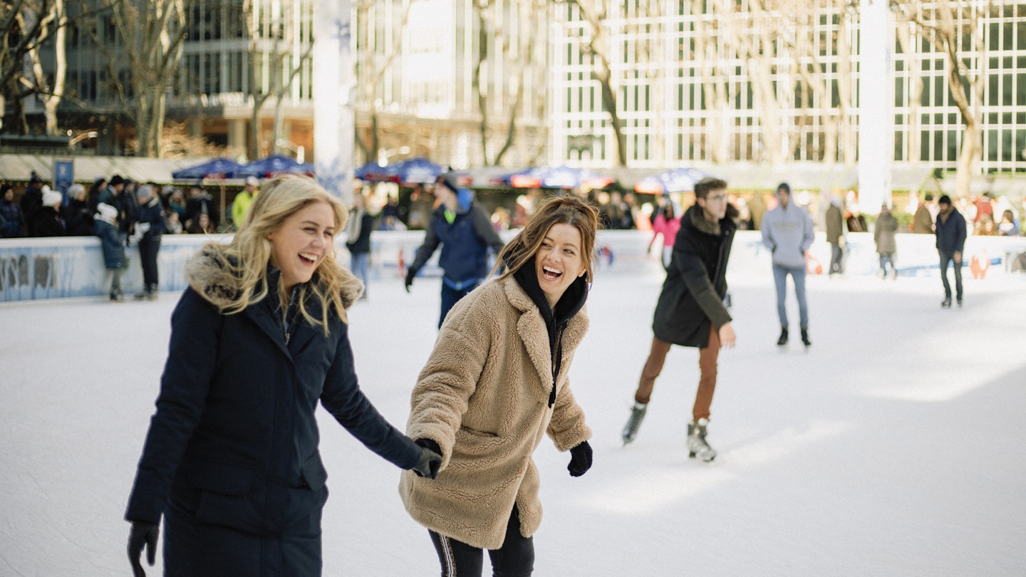 A front-view shot of two mid-adult women ice skating together in an ice rink in New York City, they are wearing warm clothing, holding hands and laughing together.