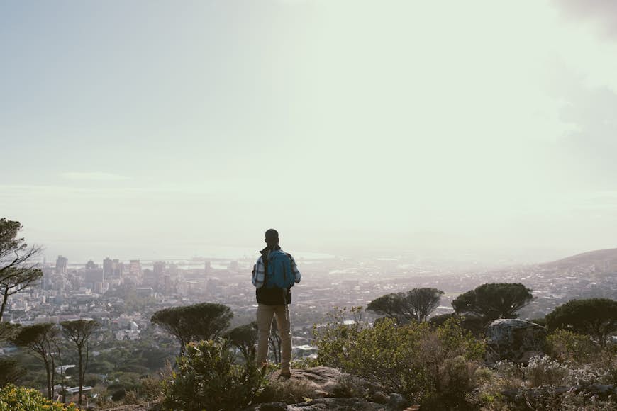 Rear view of man with standing on Table Mountain looking at the view over the city below. Full length of male hiker is on cliff by cityscape. He is with backpack against bright sky.