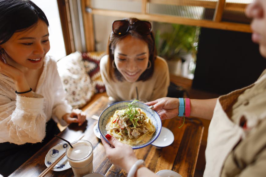Waitress bringing a bowl of noodles to a table with two customers looking excited