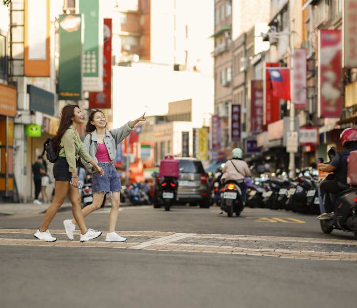 Two young Asian women come to Taipei Tamsui Old Street for independent travel in summer, with pedestrians and land motorcycle traffic in the background of old street market
