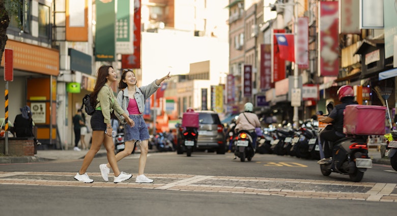 Two young Asian women come to Taipei Tamsui Old Street for independent travel in summer, with pedestrians and land motorcycle traffic in the background of old street market