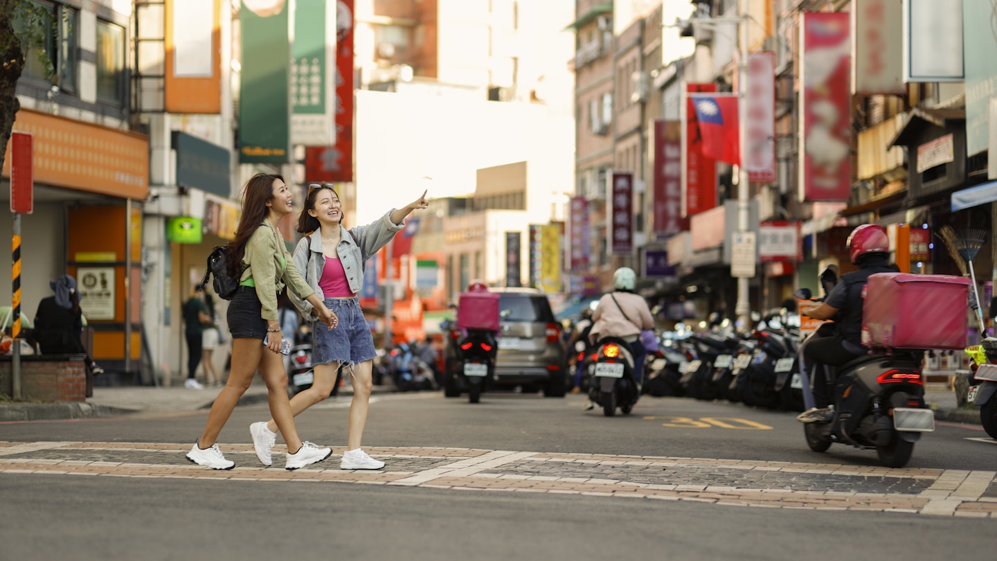 Two young Asian women come to Taipei Tamsui Old Street for independent travel in summer, with pedestrians and land motorcycle traffic in the background of old street market