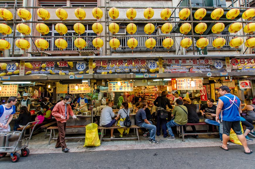 Diners sit at small tables in front of vendors cooking and selling food at a night market with yellow paper lanterns flying above them