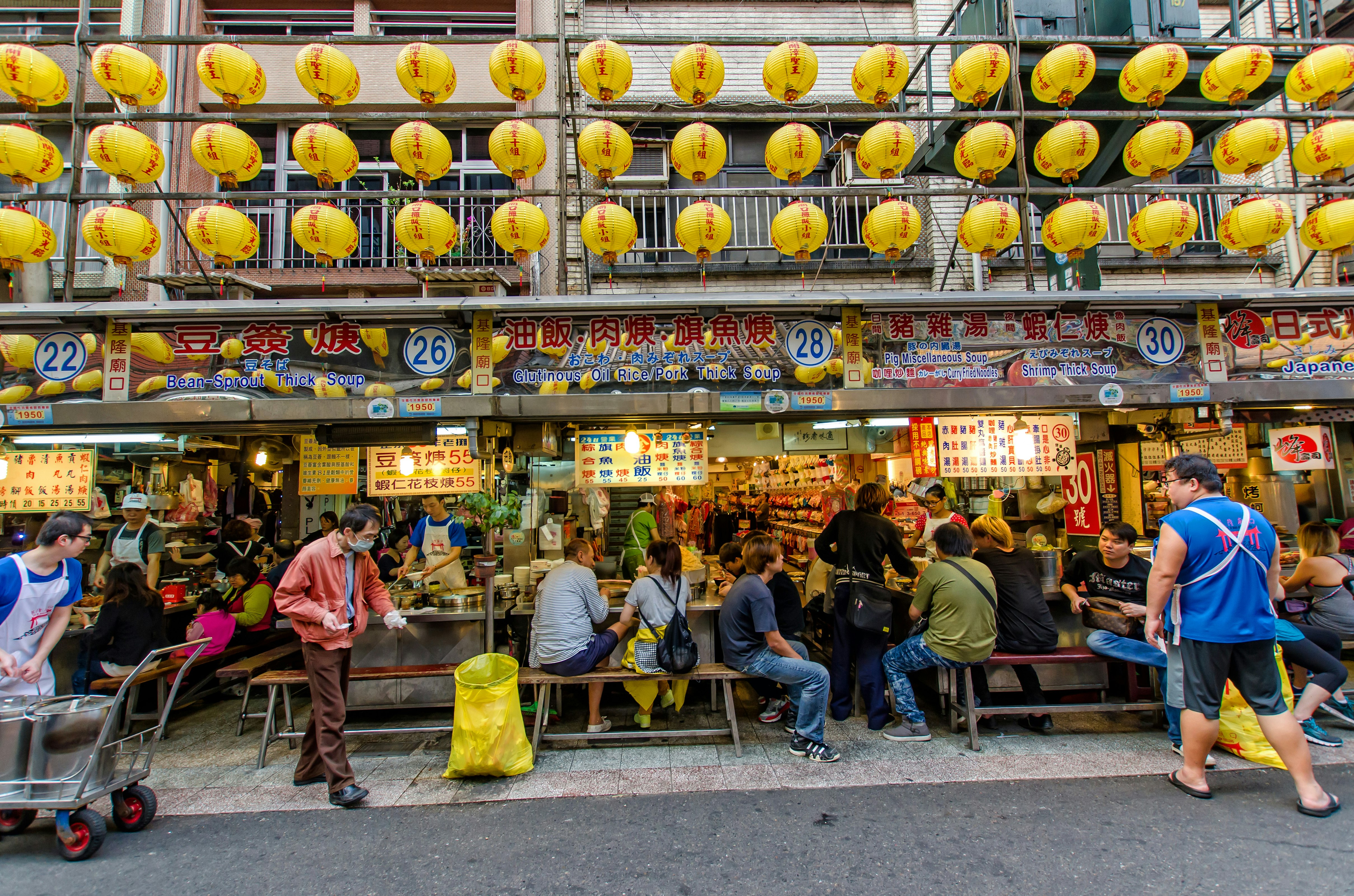 Diners sit at small tables in front of vendors cooking and selling food at a night market with yellow paper lanterns flying above them