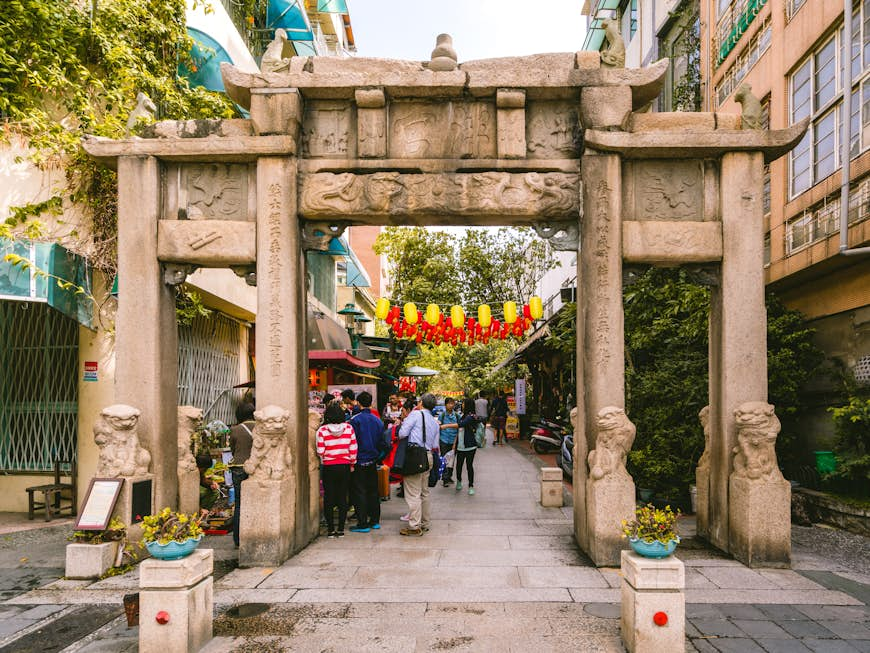 People stand near a stone gate at the entrance to a temple with yellow and red lanterns hanging above them