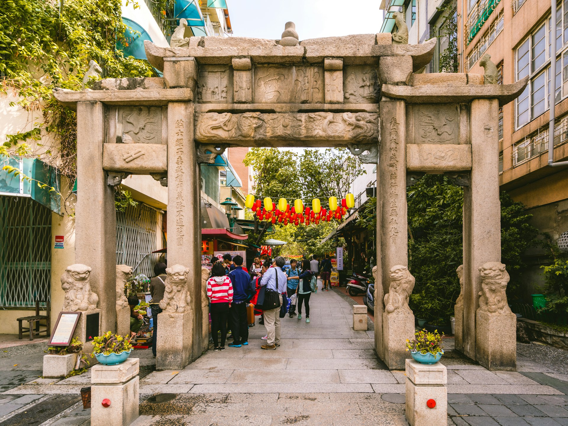 People stand near a stone gate at the entrance to a temple with yellow and red lanterns hanging above them