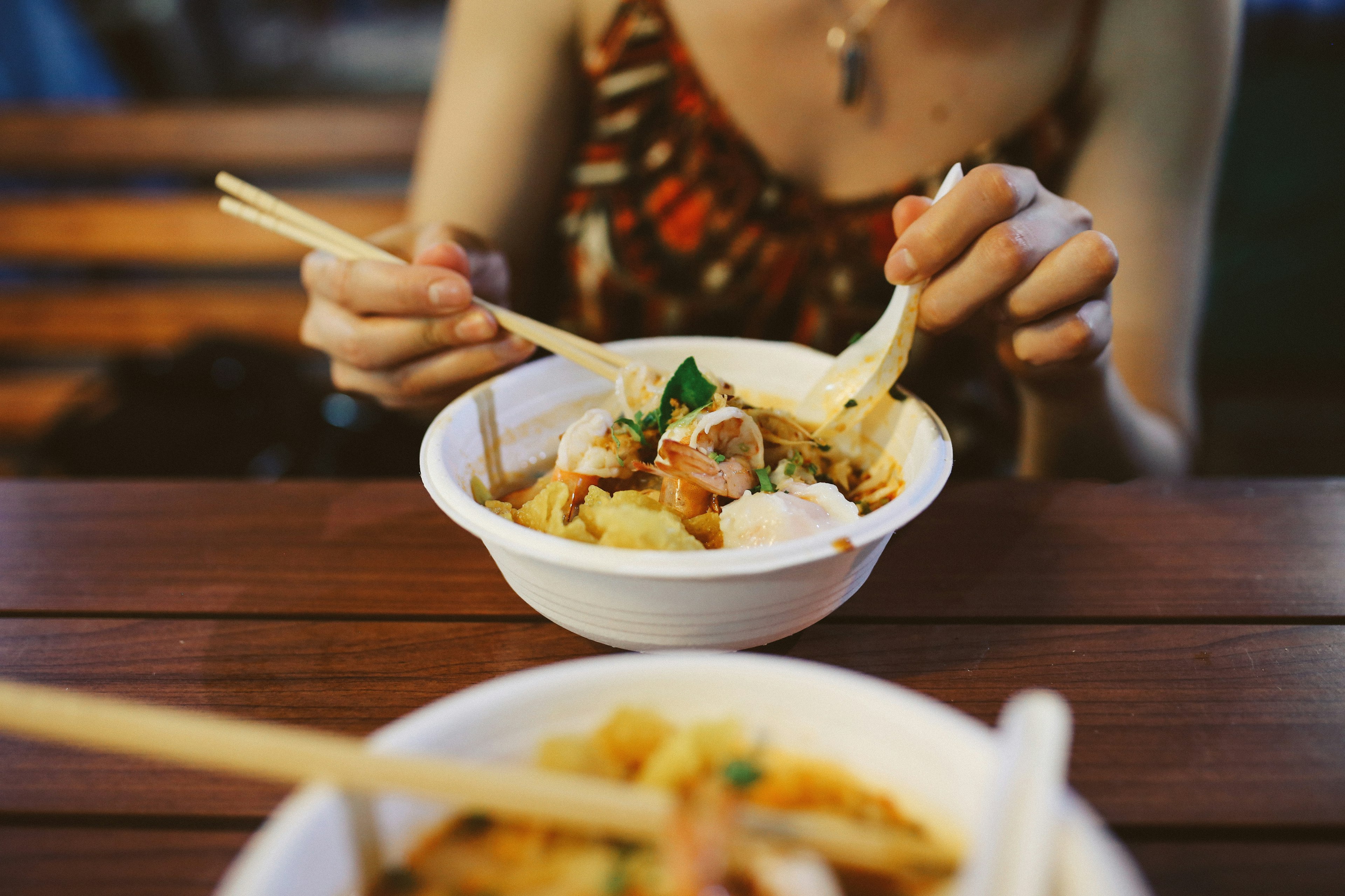 Close up of two bowls of traditional Thai food: pat tai with shrimp and red curry noodles with chicken and vegetables Served on a wooden table near Sukhumvit road in Bangkok