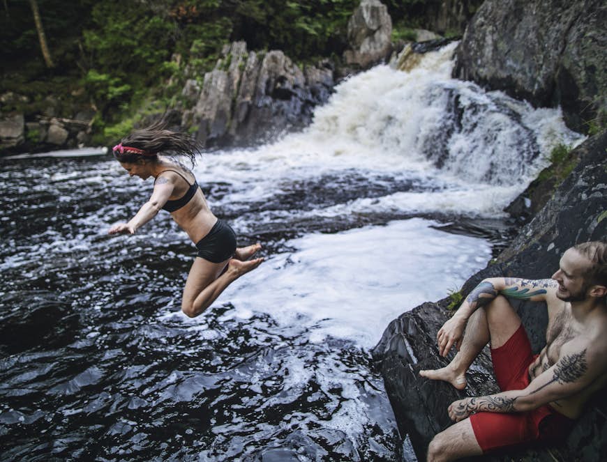 Woman smiling as she jumps into water with a waterfall thundering in the background and a man laughing as he watches her plunge