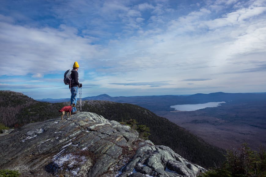 A man stands with his small dog at the summit of a mountain looking out at the view of a distant lake