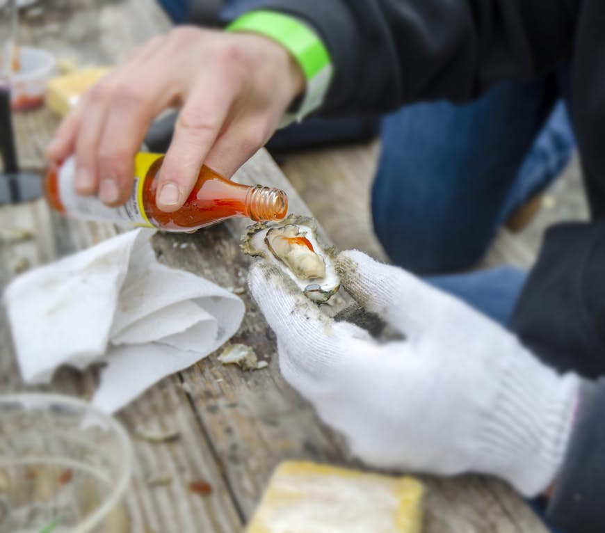A person holding an oyster pours some hot sauce onto it at the table