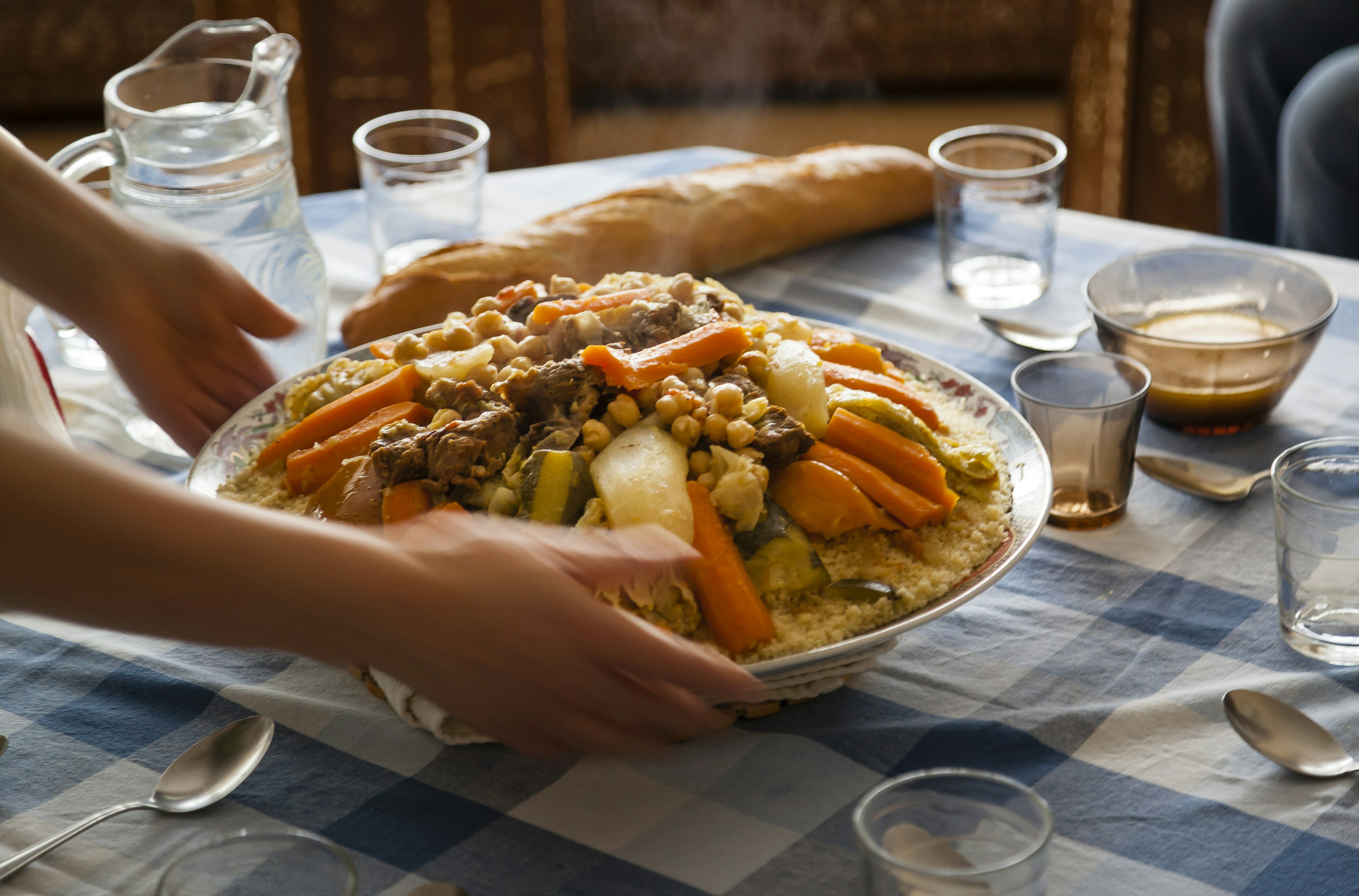 Traditional Moroccan homemade Couscous plate on a blue squared clothed table served by dynamic woman hands with glasses spoons and Laban milk .