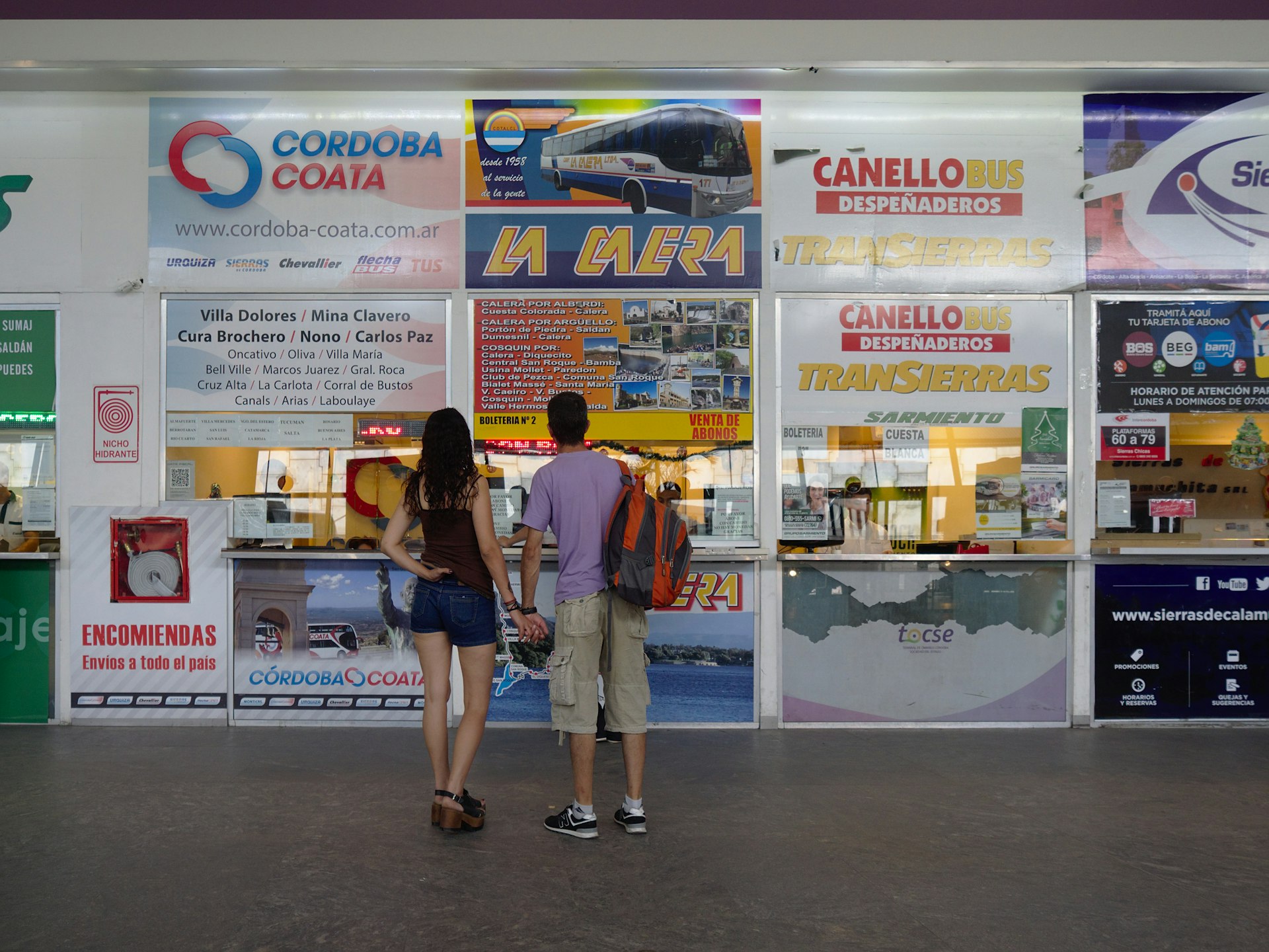 A couple stand holding hands while they look up at the ticket office displays in a bus station