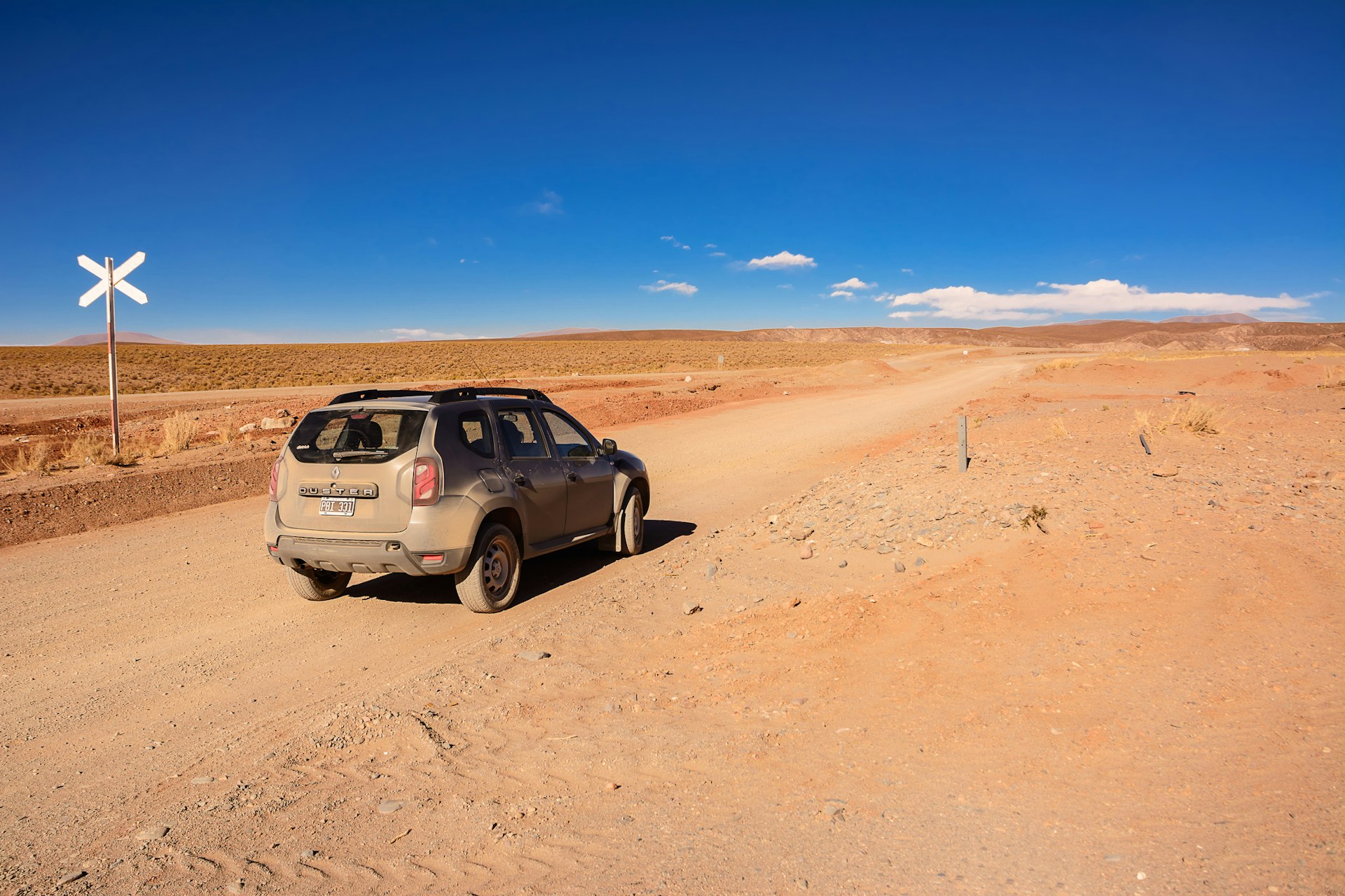 A dusty car sits on an unpaved highway surrounded by the red sands of a desert landscape