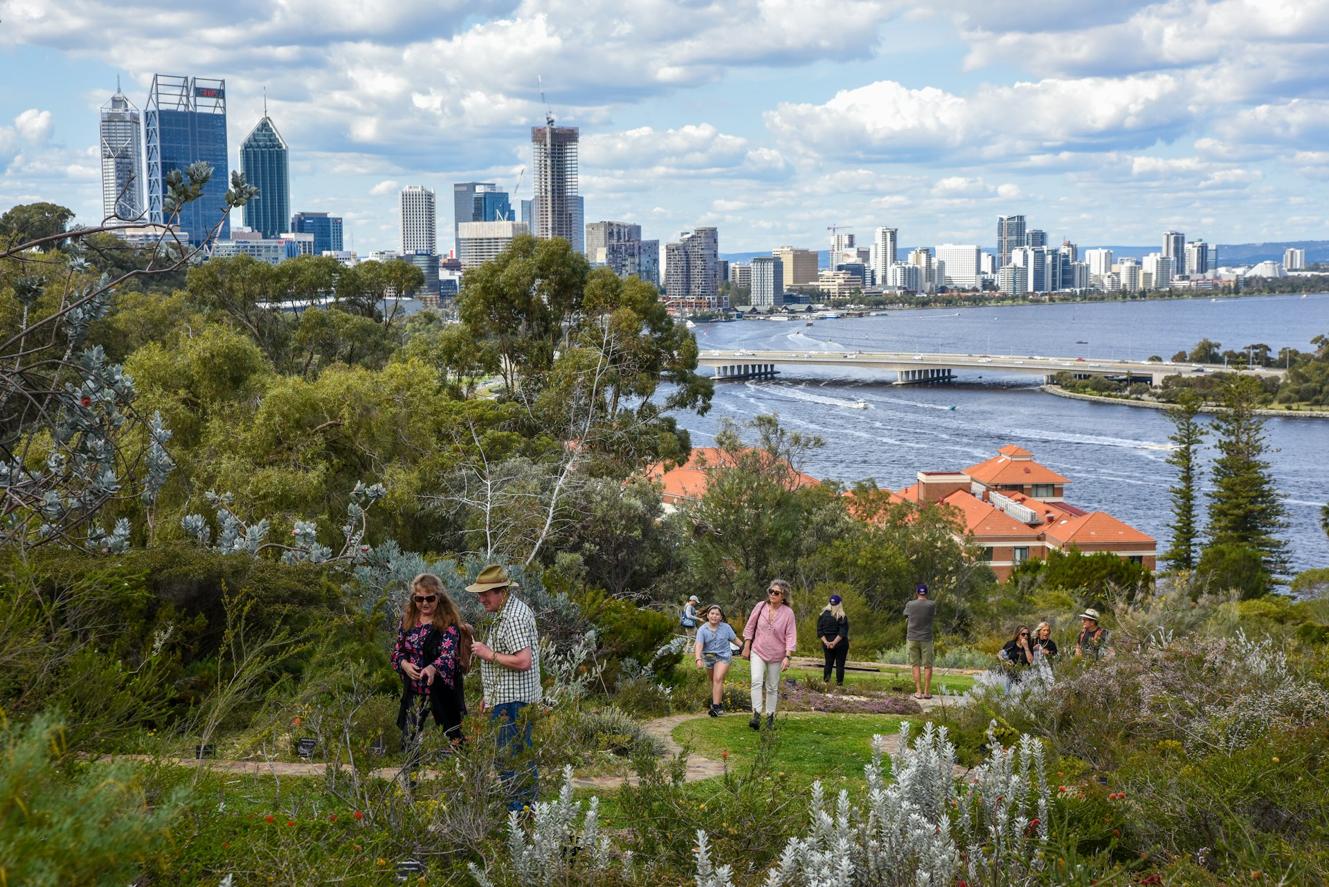 Pessoas caminham por uma trilha no exuberante Kings Park, em Perth, com a cidade ao fundo. 