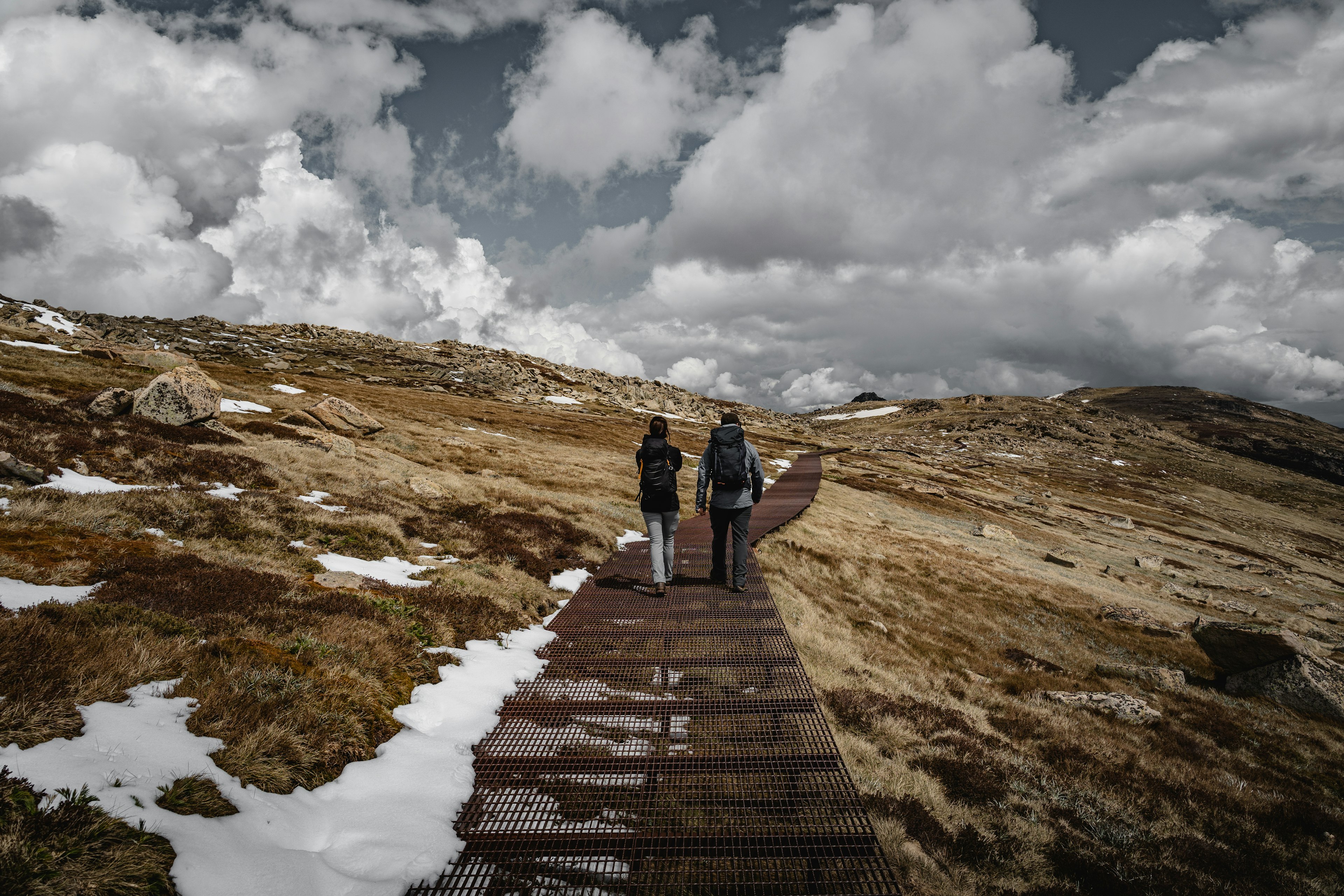 Two hikers walk along a raised trail through a snowy and hilly landscape