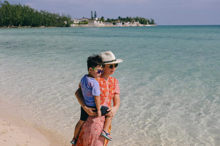 A woman holding a small child stands on a sandy beach in a tropical destination