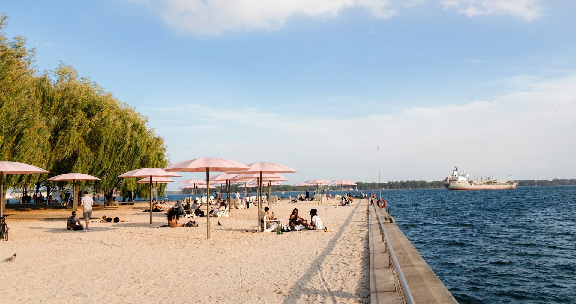 People in downtown Toronto rest under pink umbrellas on a beach beside a lake as a large cargo ship passes by