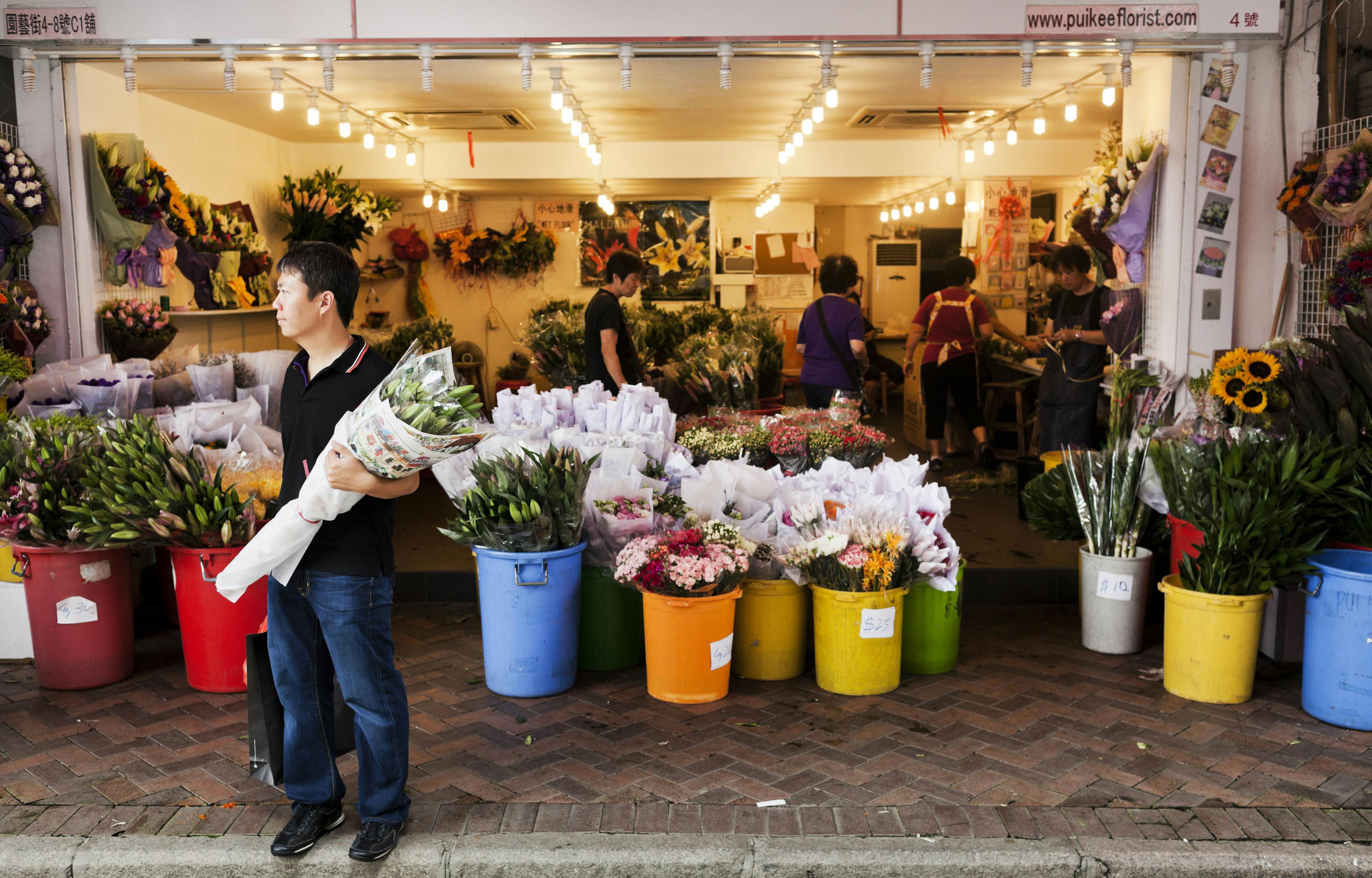 Man waiting on the street with a flower bouquet while people are shopping inside a flower shop