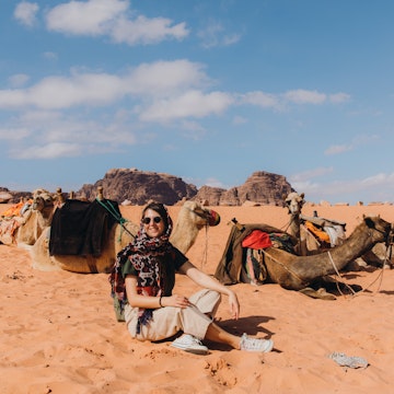 Female explorer in scarf and sunglasses relaxing at the camel bedouin camp at the beautiful landscape in the desert of Jordan