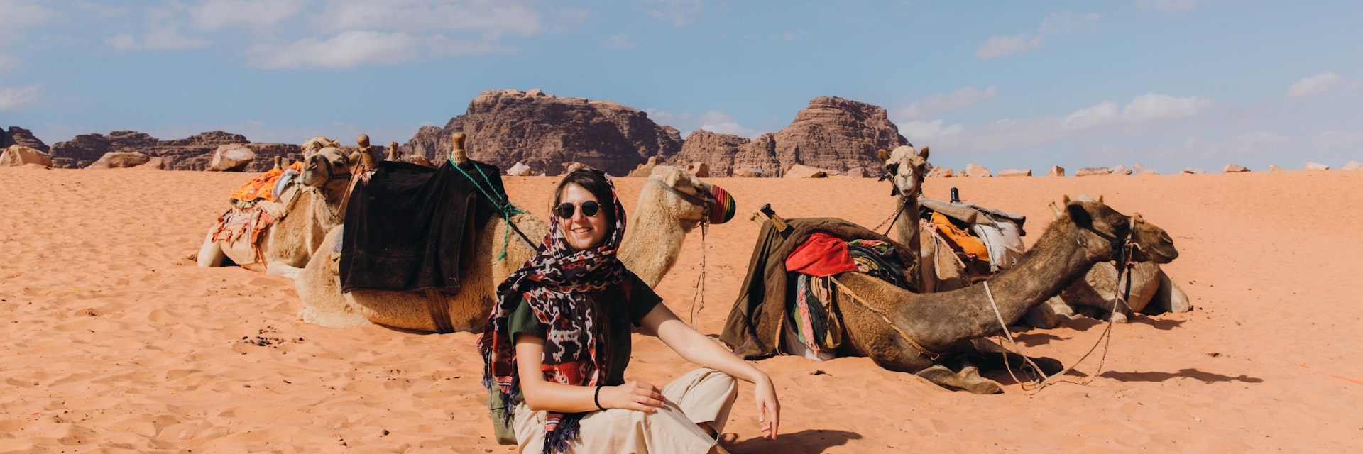 Female explorer in scarf and sunglasses relaxing at the camel bedouin camp at the beautiful landscape in the desert of Jordan