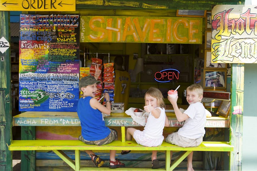 Kids smile at the camera while sitting at a shaved ice stand. 