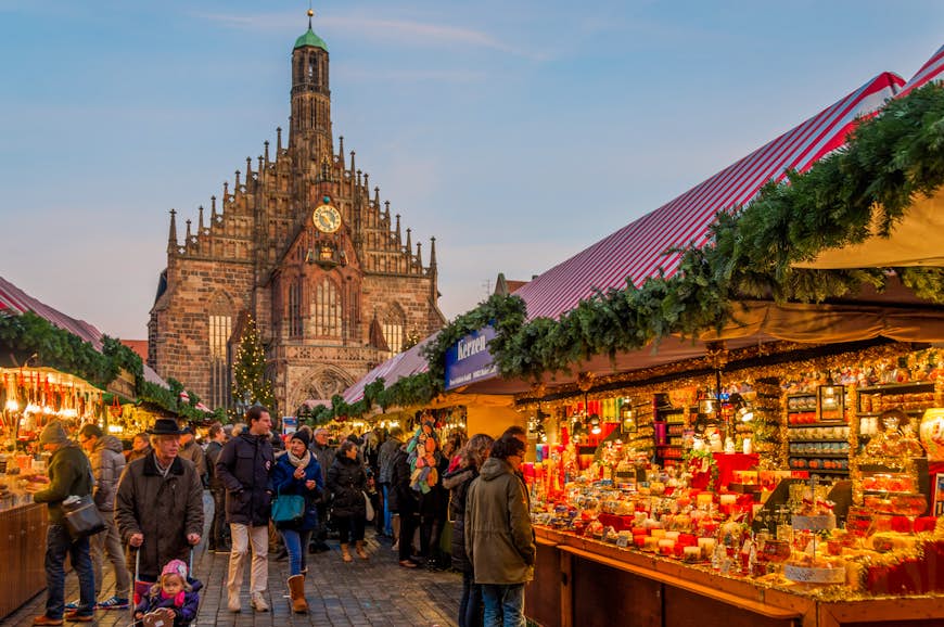People walk through a lit up Christmas market in front of an elaborate church. 
