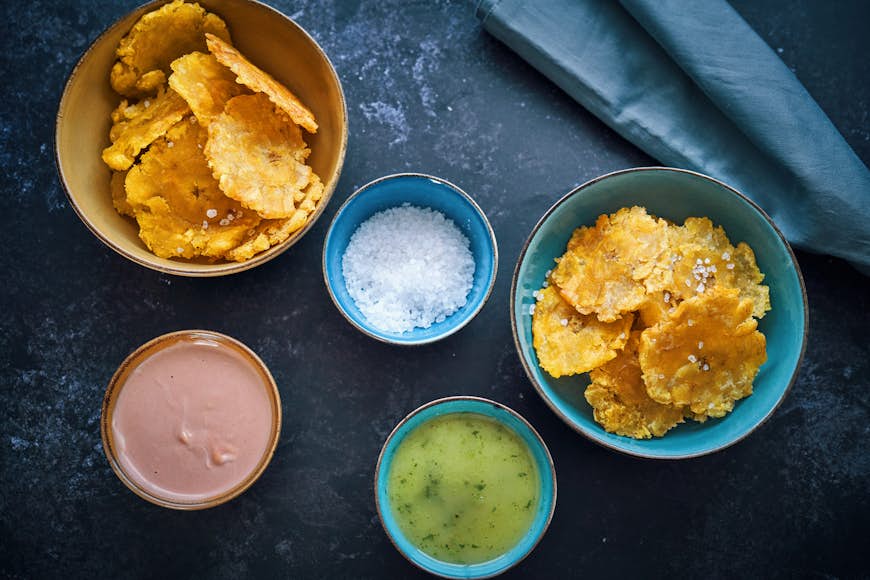 Fried plantain dishes with colorful dips of pink and green, alongside a pot of salt