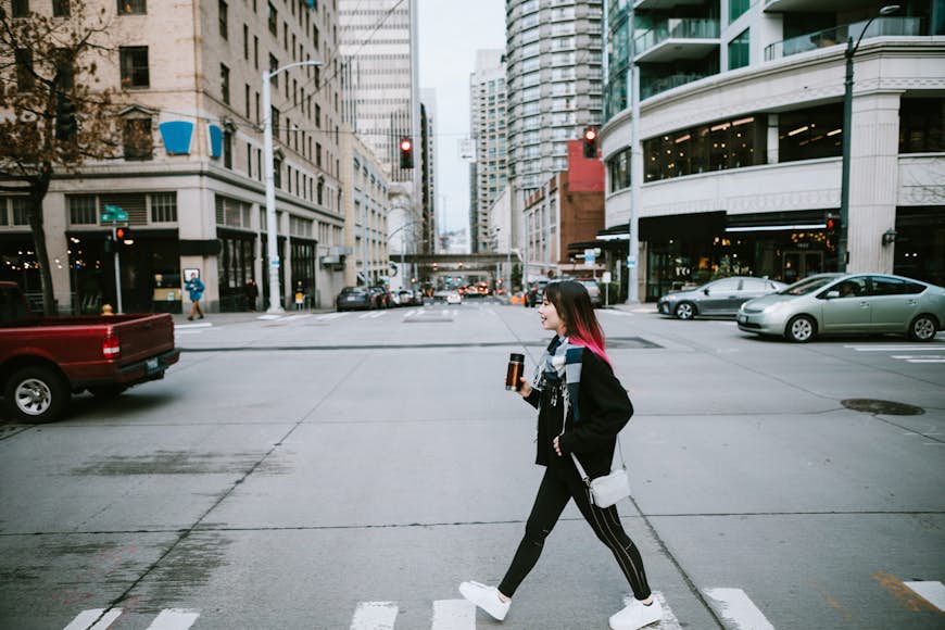 A young woman enjoys time in the city of Seattle, Washington.  She walks the streets, holding a reusable coffee container.