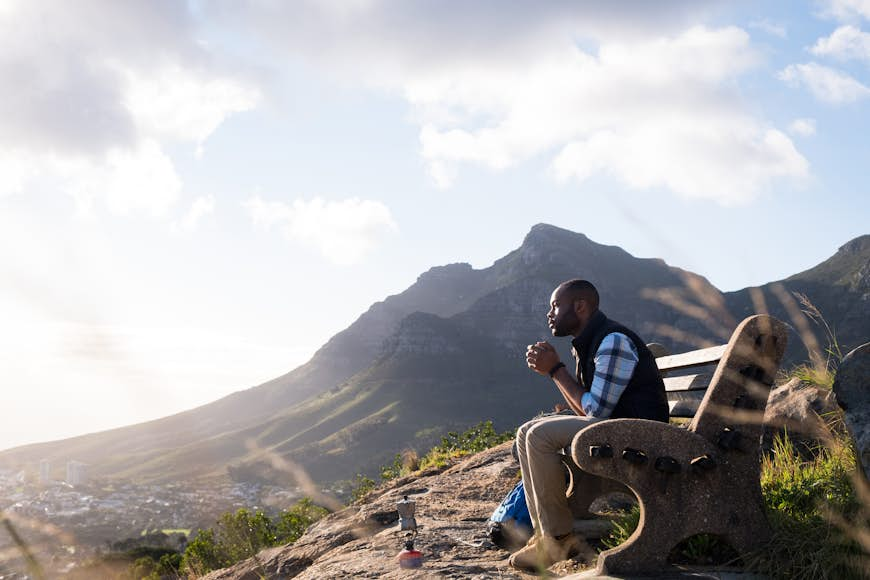 Side view of male hiker sitting on a bench, having a drink and looking at Table Mountain. 