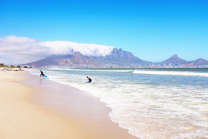 Surfers wearing wetsuits emerging from the Atlantic Ocean at Milnerton, north of Cape Town, on a hot summer's day. Behind them is the city center and the world-famous landmark of Table Mountain.