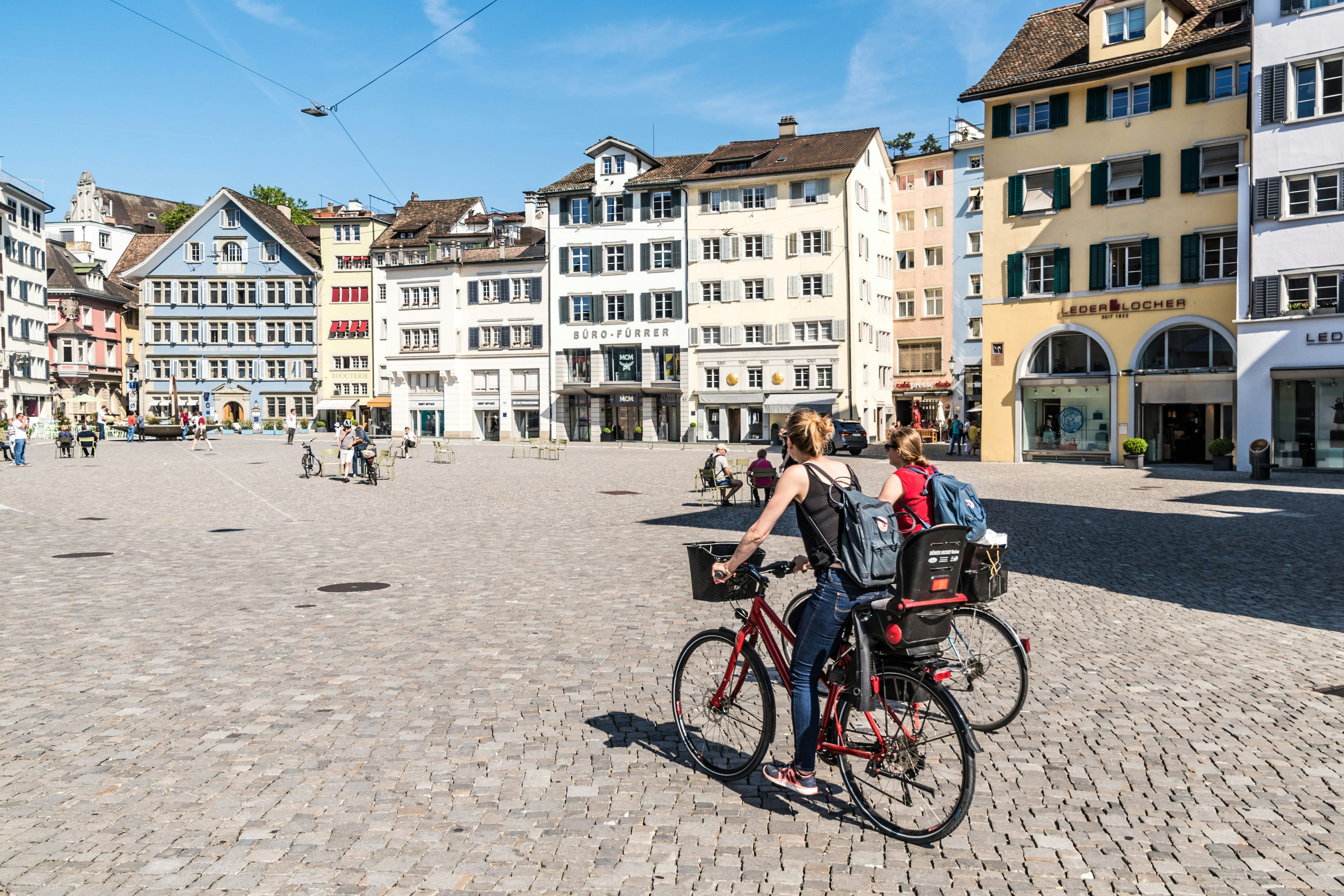 Two yound ladies ride bicycles through a town square in Zurich, the capital of Switzerland. Other people walk along the paths passing shops.
693239720