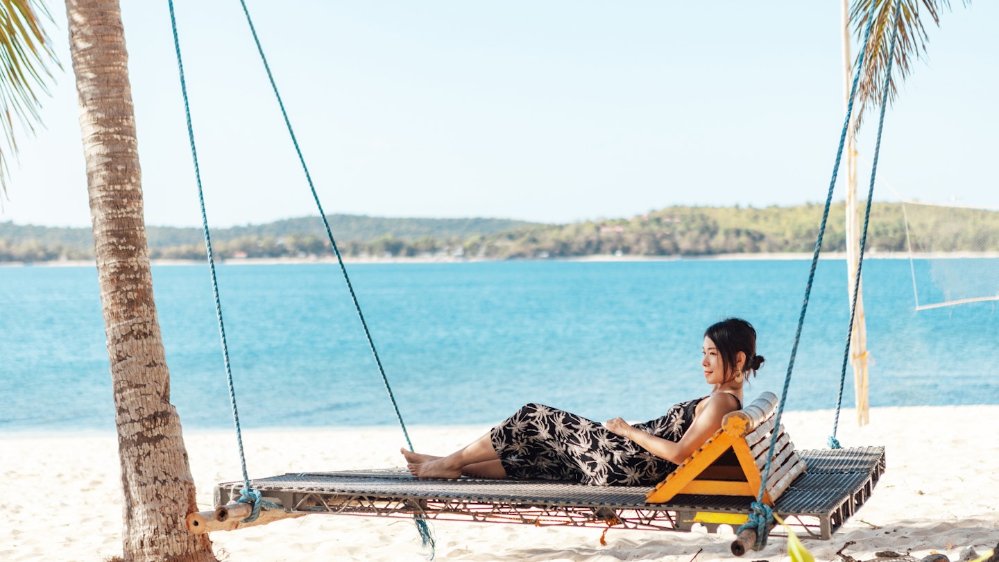 Young beautiful Asian woman looking at view while sitting on hanging bed near the beach. Tropical beach holiday. Local experience travel. Wanderlust. Summer vacation.