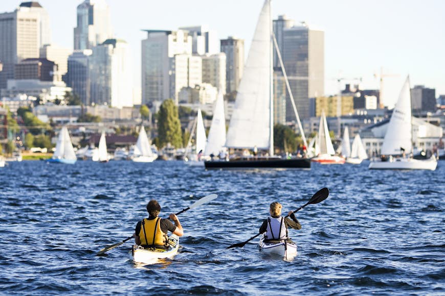 Young man and woman each paddling a kayak on blue lake, cityscape with sailboats in background