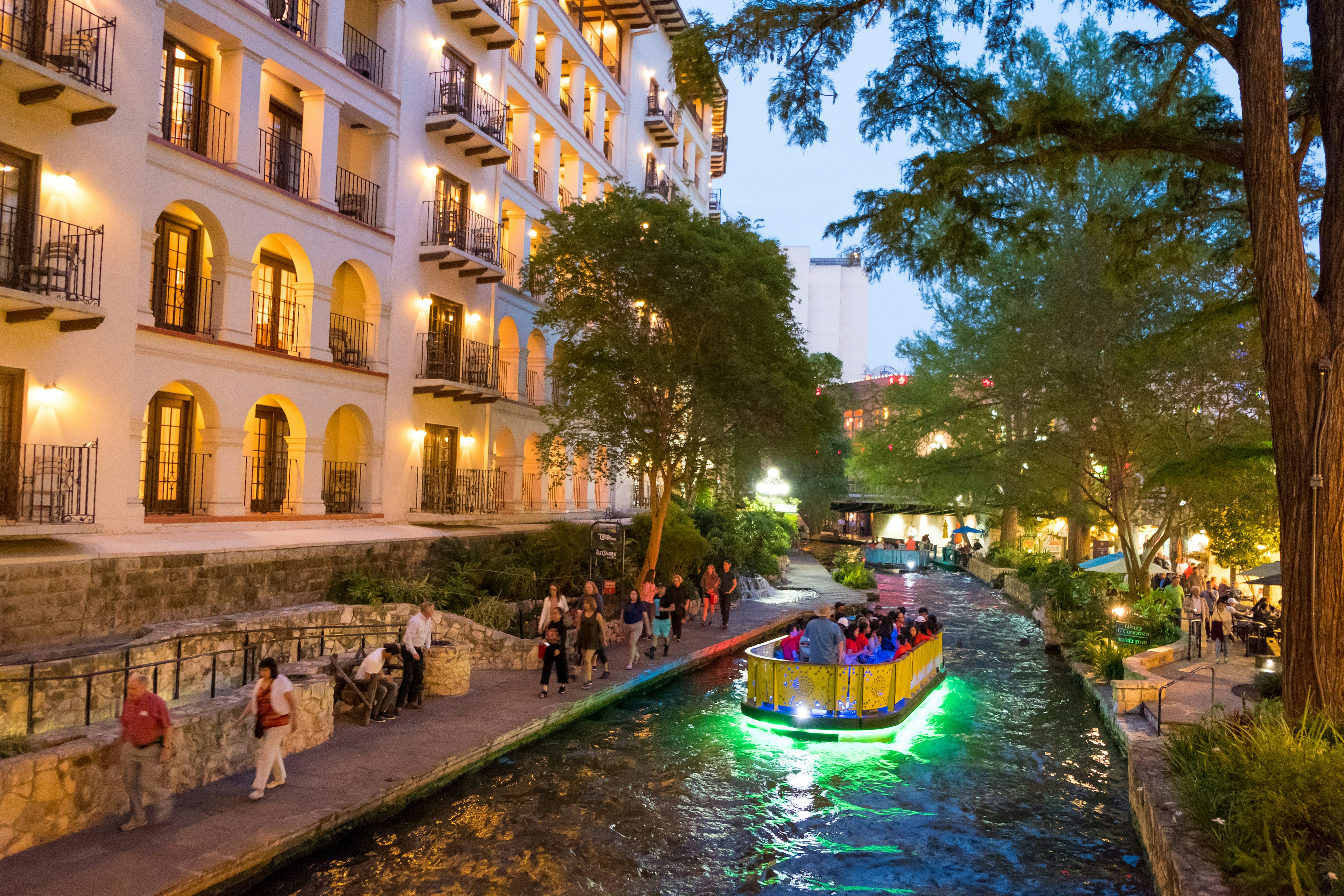 A boat, with a hull lit up in green, floats down a river in a city centre at twilight