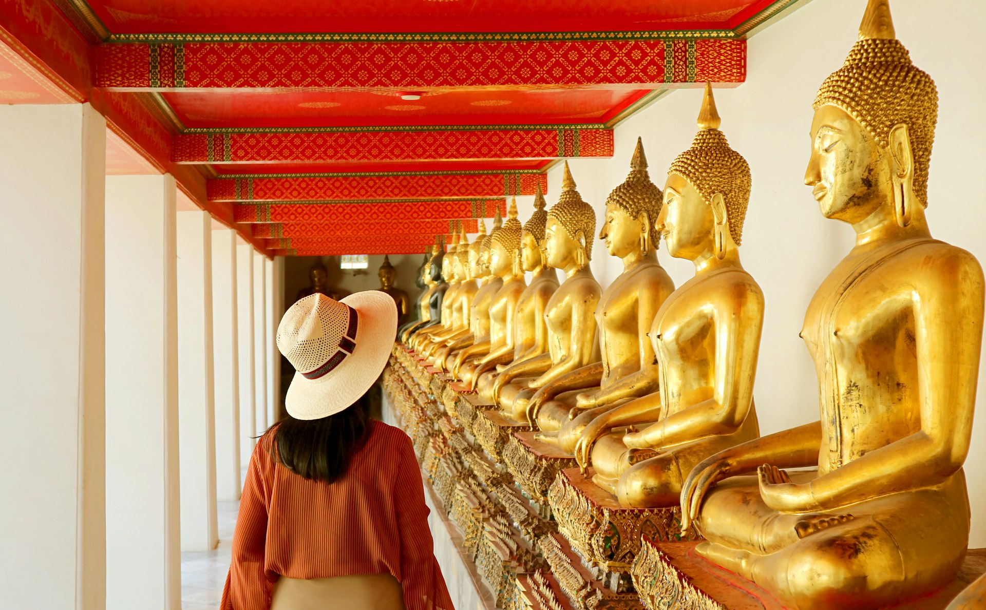 A woman wearing clothes that cover her shoulders walks along a wall of golden buddhas in a Thai temple