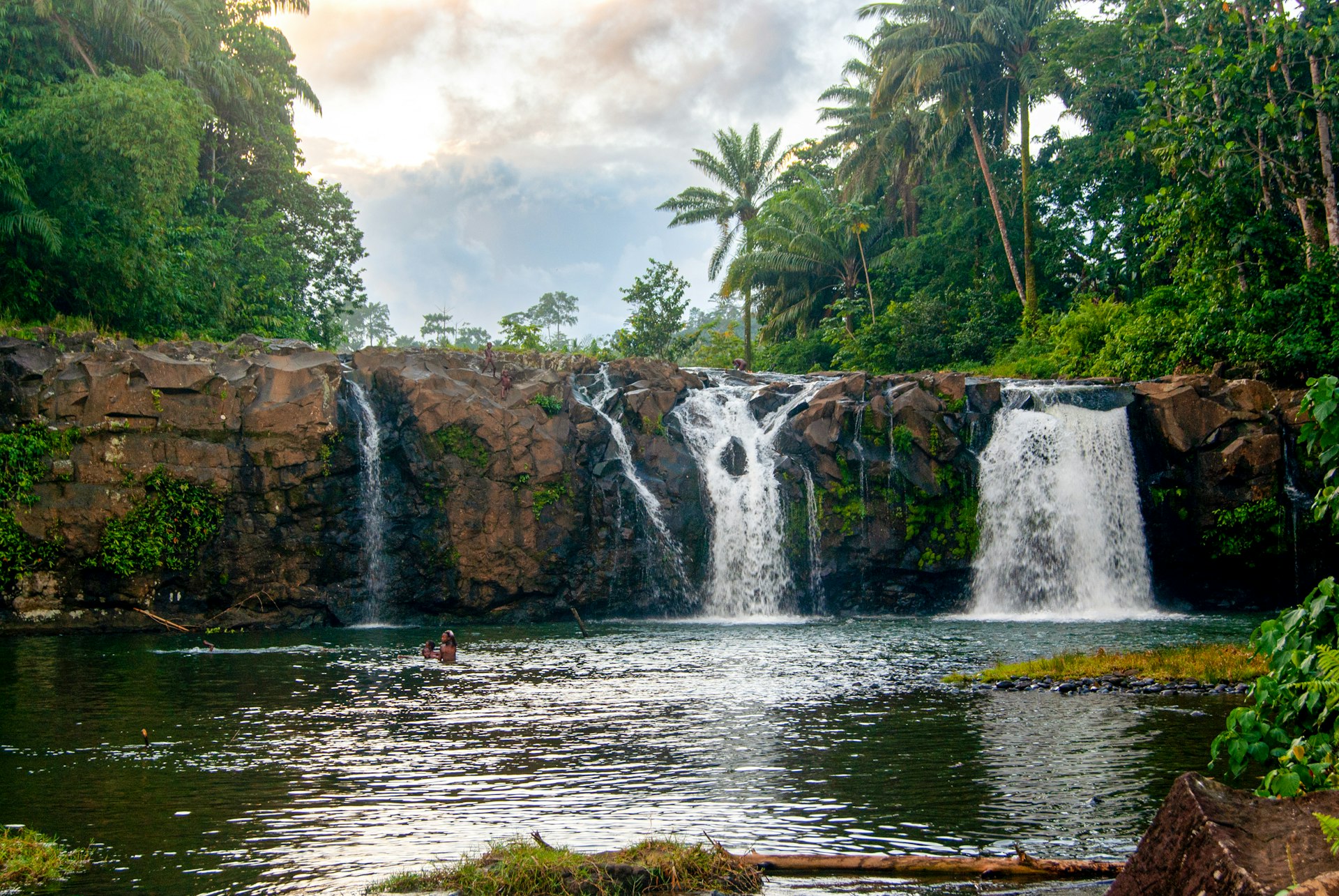 People swim below a beautiful waterfall in a lush forest. 