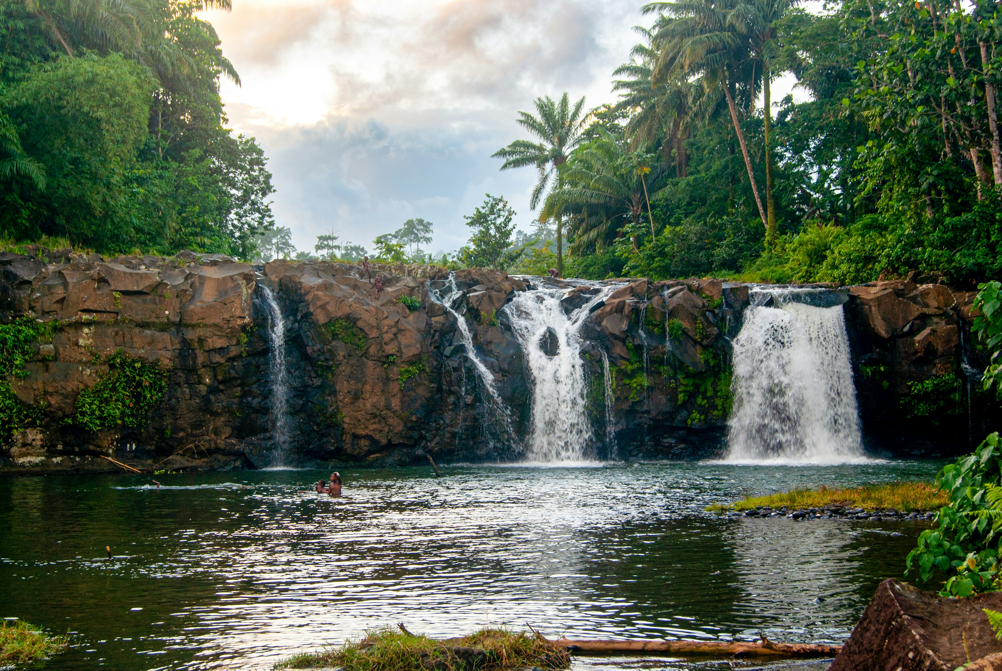 People swim below a beautiful waterfall in a lush forest.