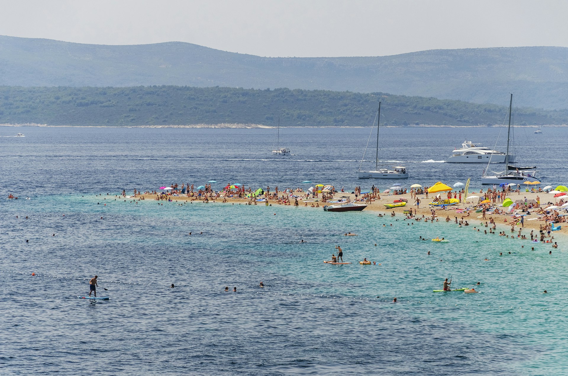 Many people crowd onto a beach spit in Croatia while others play or paddleboard in the sea