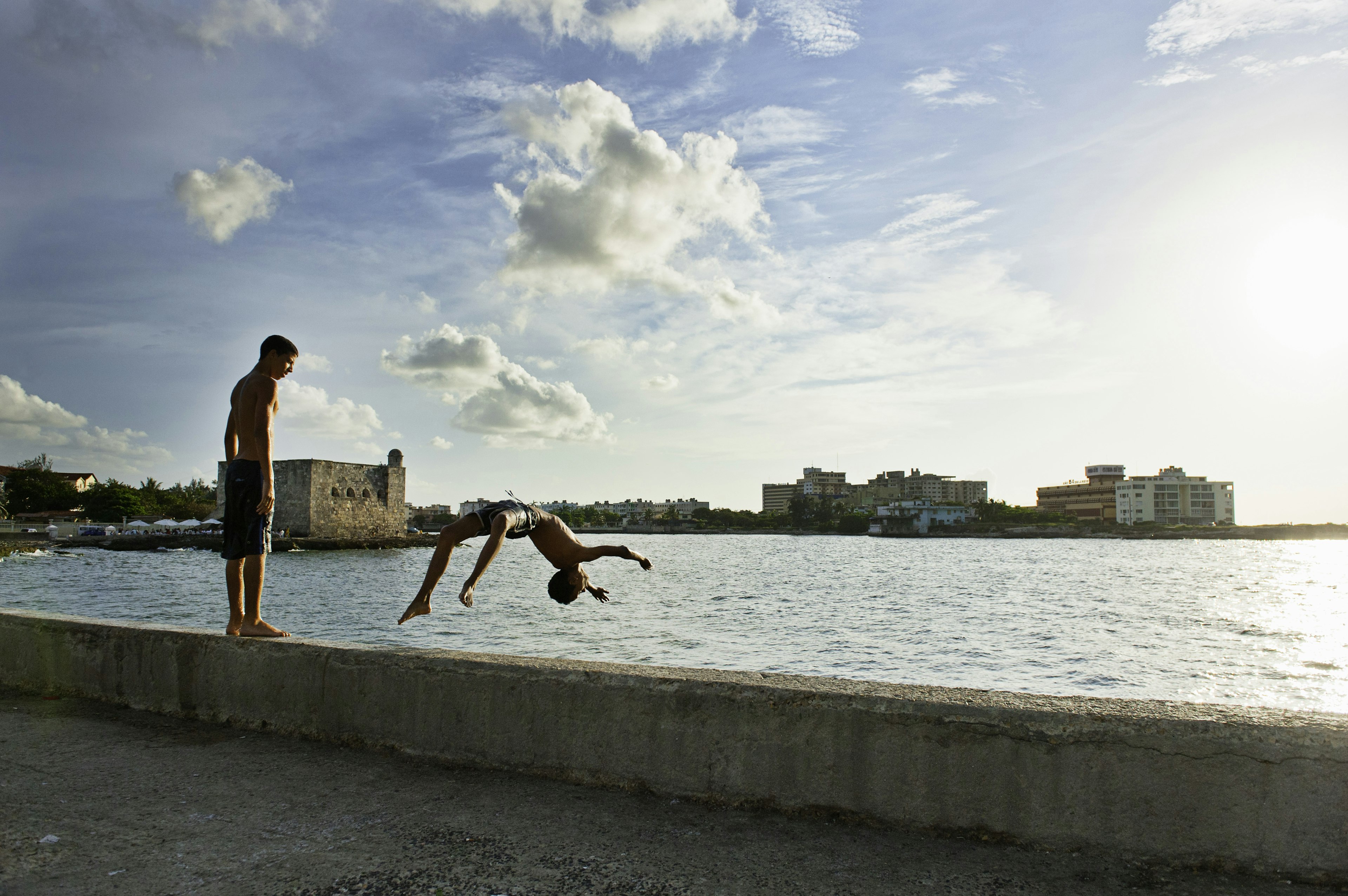 A boy stands on the harbor wall in Havana, watching another boy backflip off into the water below