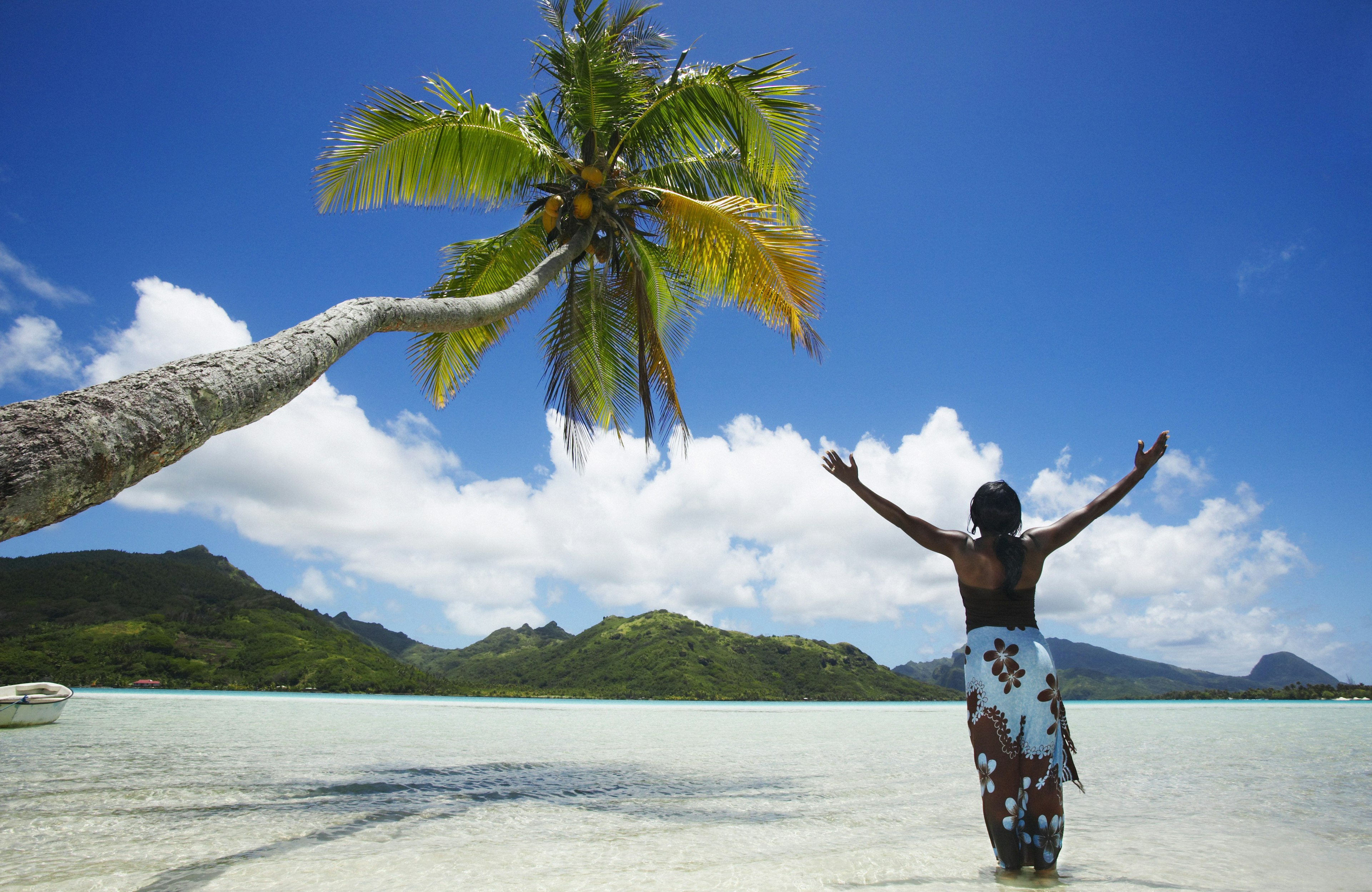 A woman stands with her arms raised to the sky on an idyllic beach in Tahiti.