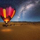 A majestic hot air balloon soars under the stars of the Milky Way, over the desert - Mesquite Dunes of Death Valley National Park. Moonlight provides luminosity showing the patterns and shapes of the desert landscape.