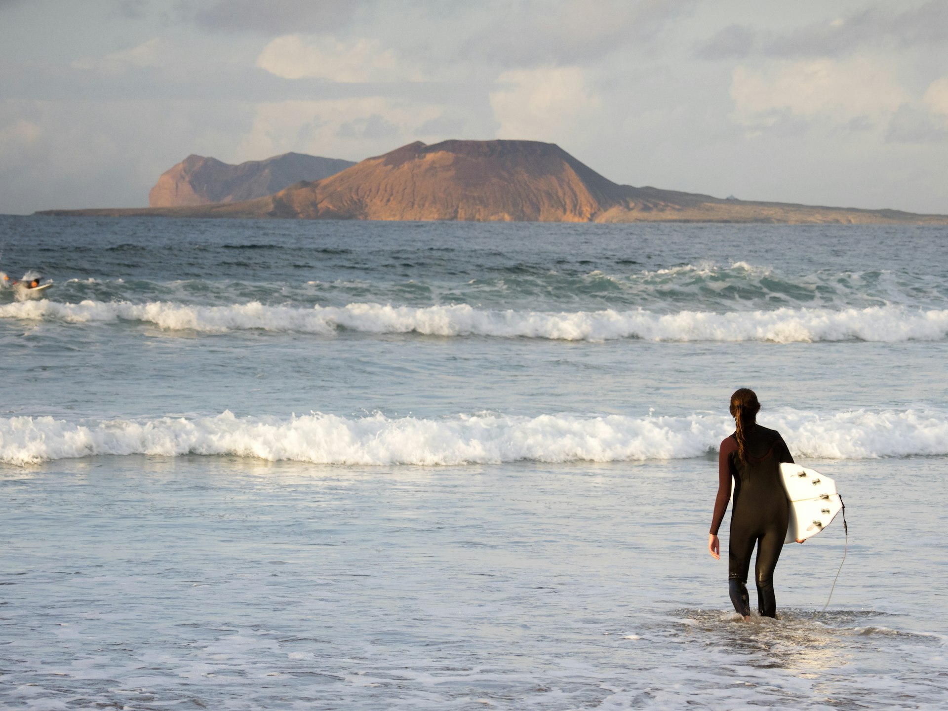 Surfer walks into the water at the beach of Famara, with the La Graciosa Island in the background, Lanzarote, Canary Islands, Spain