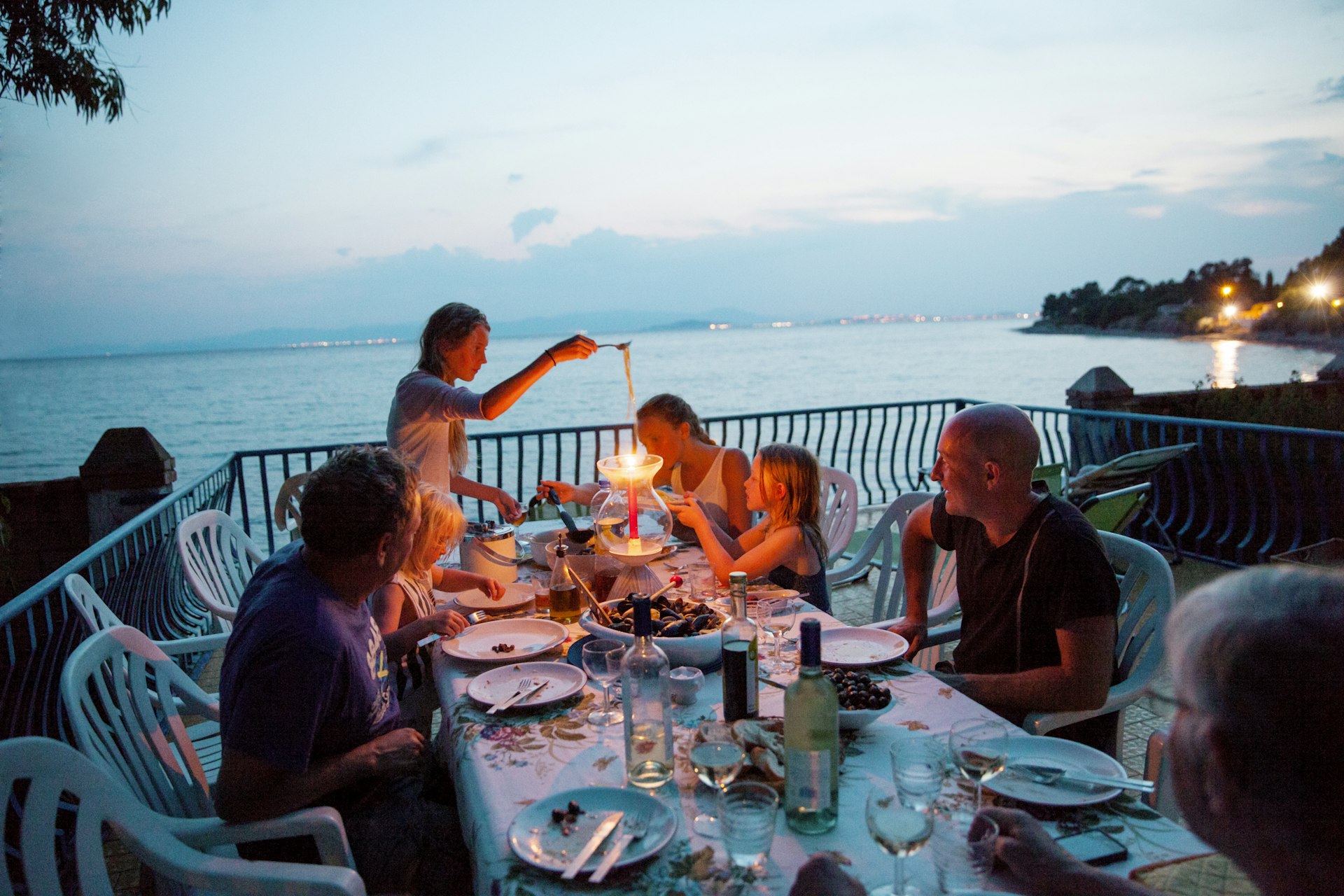 A family with several children ranging in ages and adults eat together at a table next to the sea