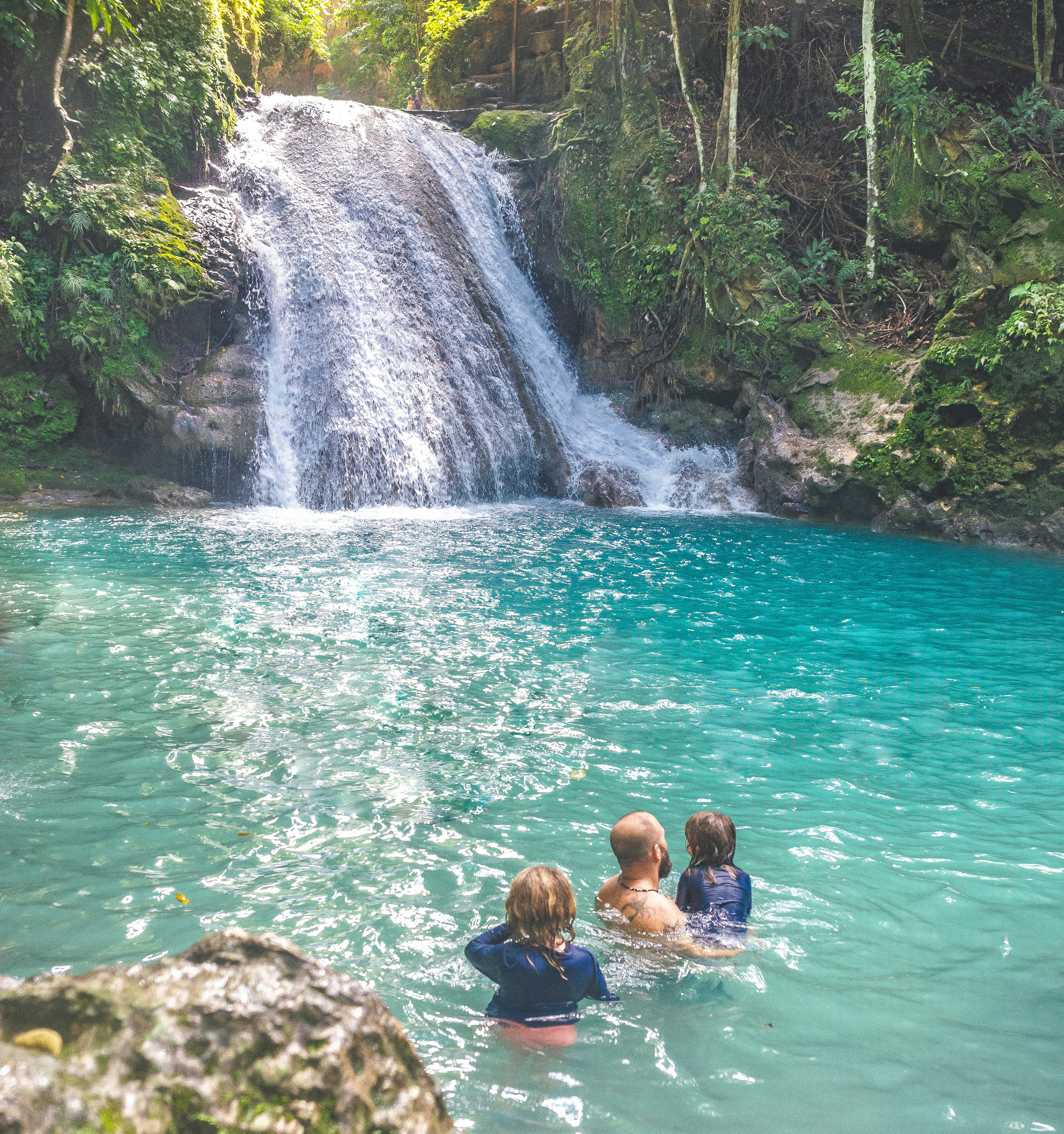 A man and two children swim in a pool at the foot of a waterfall surrounded by jungle